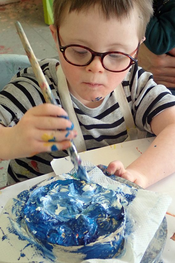 young boy with Down's Syndrome painting with blue and white paint. The boy has blue paint on his chin.