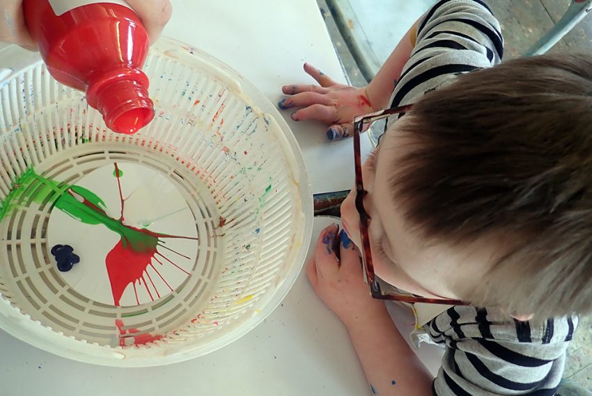 a birdseye view of a you boy with a hand pouring red paint into a colander to do a splatter paint picture.