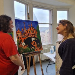 A man and a woman stand in a studio. She is volunteering at the studio. There is an artwork in an easel on the background.