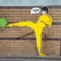 graffeti artwork on a wall of a man dressed in a yellow tracksuit kicking a symbol of coronavirus