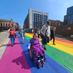 A group of people walking and wheeling through a pride rainbow painted pavement.