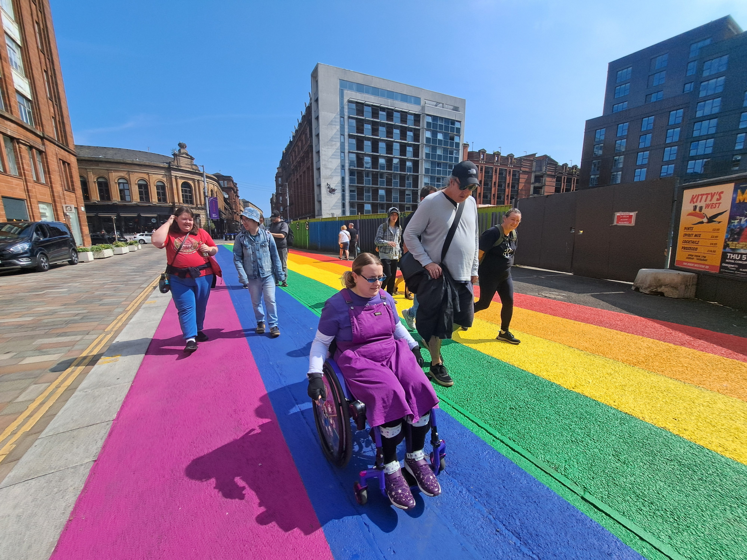 A group of people walking and wheeling through a pride rainbow painted pavement.
