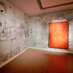 An image showing two walls of the gallery. You can see the red wooden piece hanging to the right of the back wall. the rest of the walls are covered in etchings and marks.