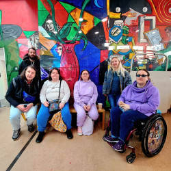6 members of the walking group are standing or sitting in front or a large colourful abstract painted wall to get their picture taken