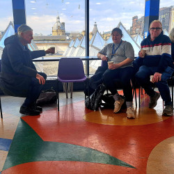 three people from the walking group are sitting on blue chairs drinking coffee in front of a large window.