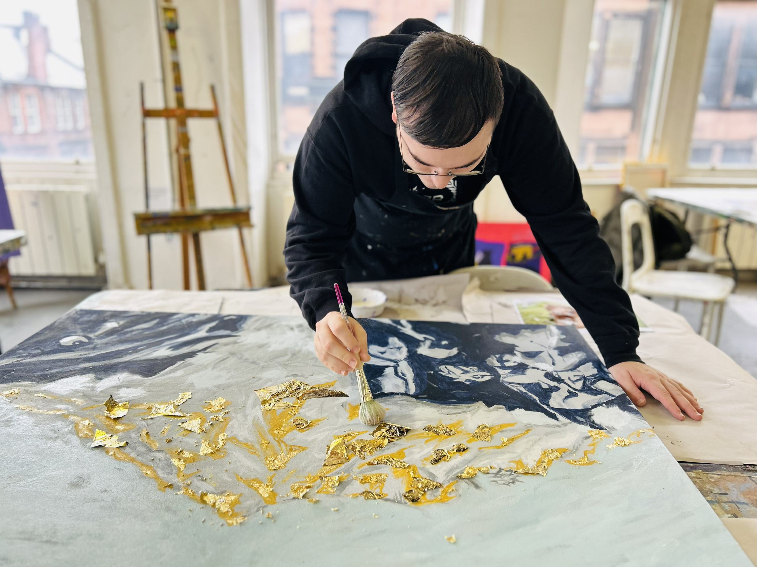 an artist leaning over a painting on a table. he is holding a paintbrush and painting on gold leaf
