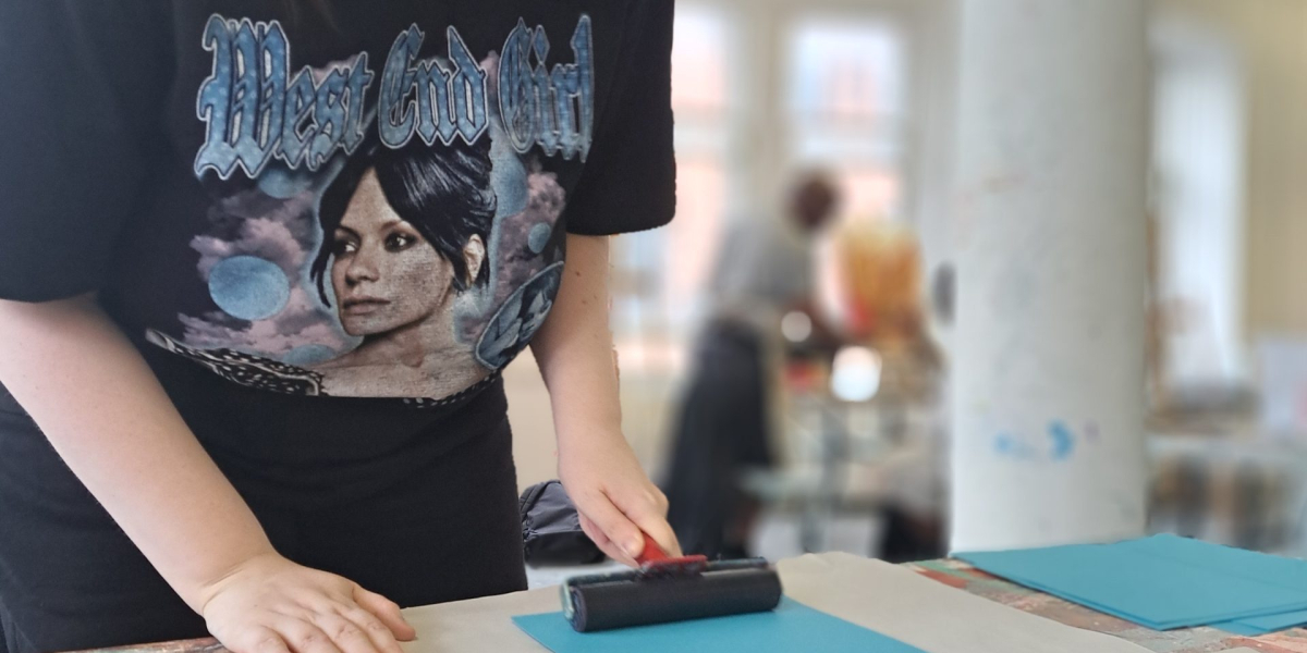 Woman wearing glasses and a graphic T-shirt carefully presses a print onto blue paper at an art table in a bright studio, focused on her craft.