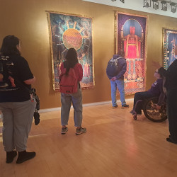 Visitors gather in a warmly lit gallery to view vibrant tapestries, including a sun motif and robed figure; a guide speaks to the group.