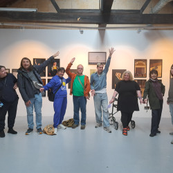 Diverse group of nine people pose in an art gallery, smiling and raising arms
