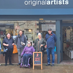 A group of six people stand outside the originalartists shop, with one person in a wheelchair next to a sign for a Project Ability art exhibition.