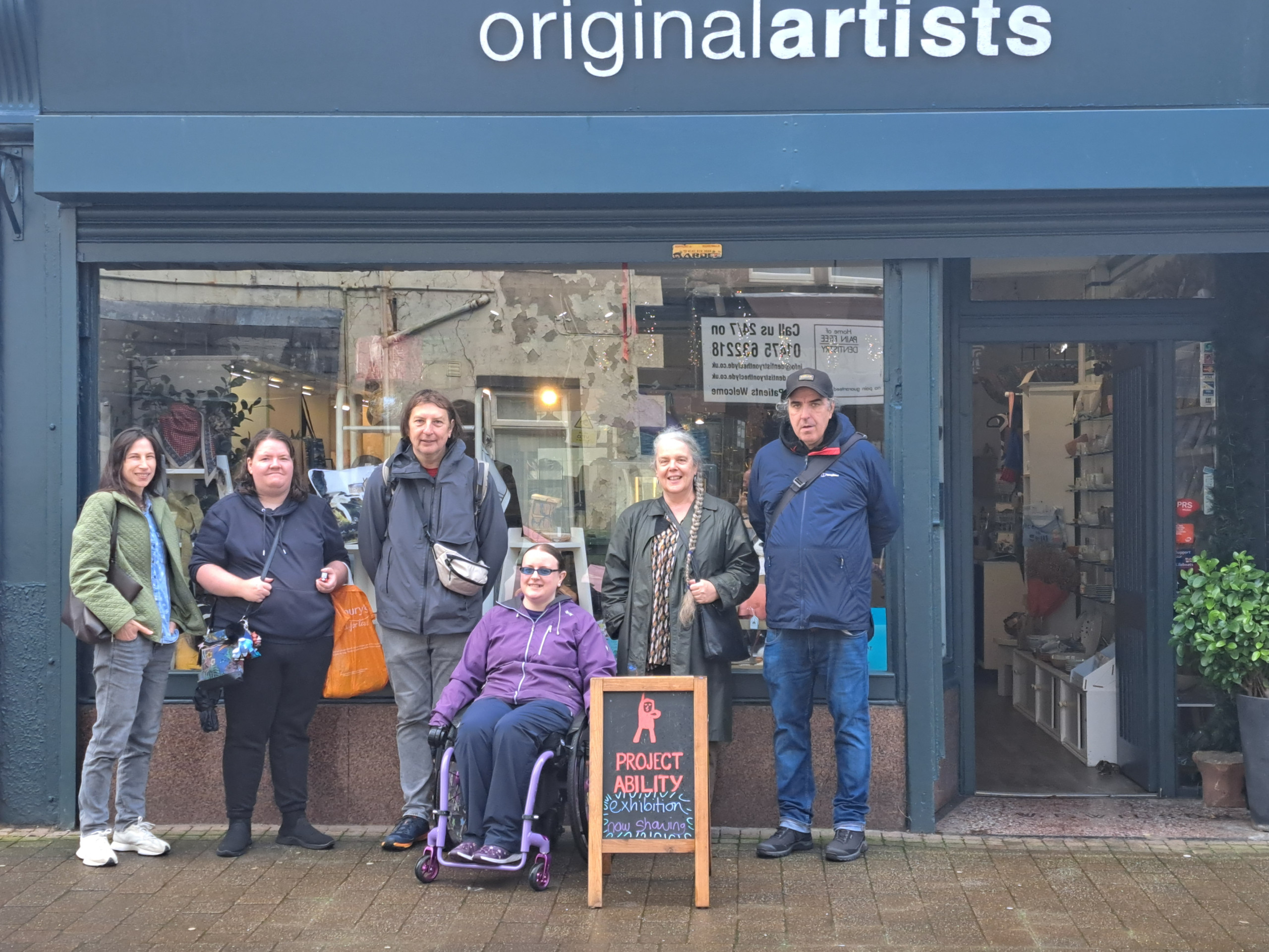 A group of six people stand outside the originalartists shop, with one person in a wheelchair next to a sign for a Project Ability art exhibition.