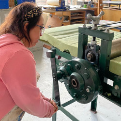 A woman in a pink sweater operates a large green printing press in a bright art studio with tall windows.
