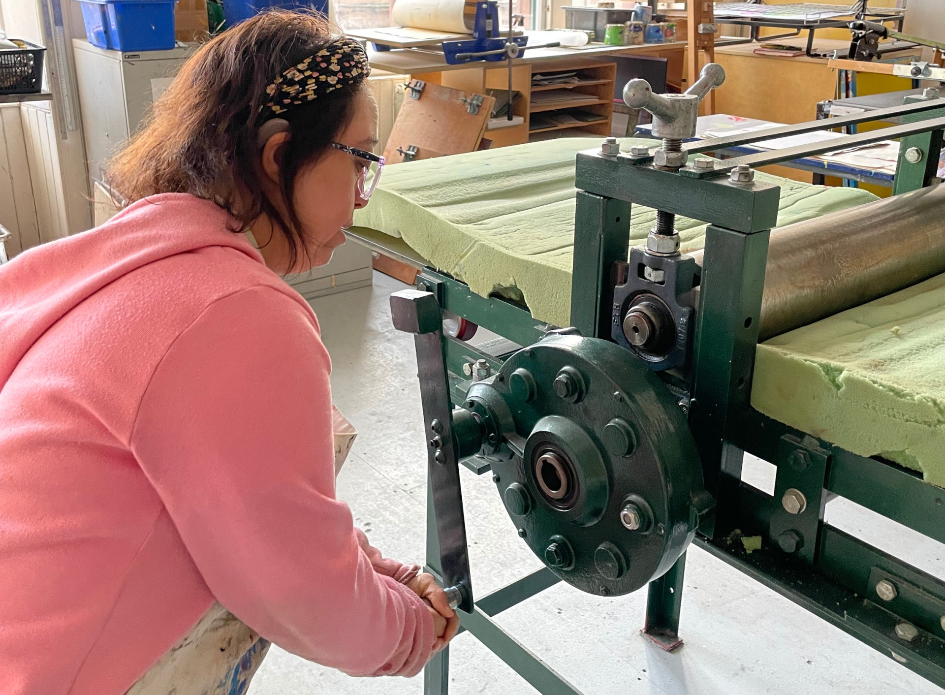A woman in a pink sweater operates a large green printing press in a bright art studio with tall windows.