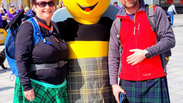 Vikkie and Iain posing for a photo with a bee mascot wearing a kilt.