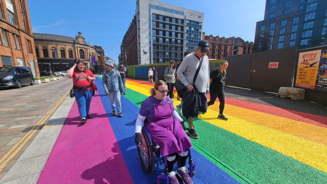 A group of people walking and wheeling through a pride rainbow painted pavement.
