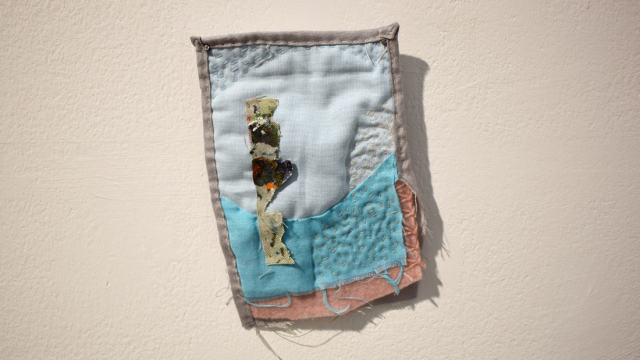 A small quilted piece with visible stitching. Collaged fabric pieces in light blue, a sky blue, a soft pink and a soft grey. A wiggly column is stitched onto the piece in a fabric covered with paint.