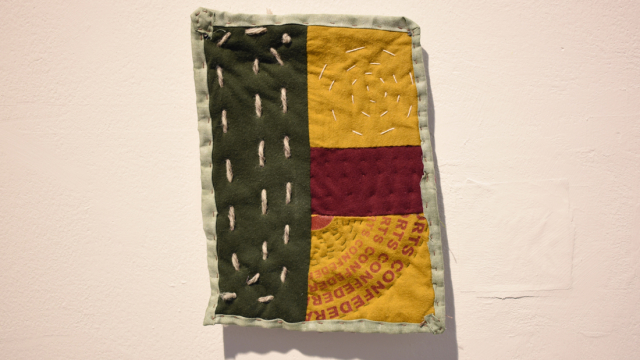 A small quilted piece with visible stitching and collaged fabrics in crimson, ochre and dark green with a light green border. One patch of fabric has been repurposed from something with printed red words on it.