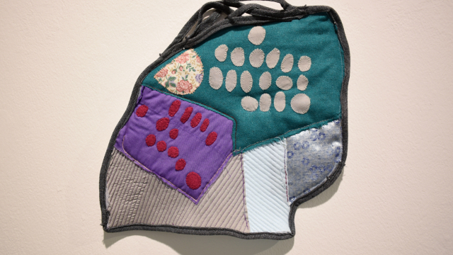 A small irregular shaped quilted piece with embroidered and appliqued oval shapes stitched onto a collaged fabric background and bordered in a black denim fabric.