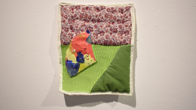 A close up of a small wall quilt with a cream border. A flowery fabric and two green fabrics are collaged together with a piece of joyful painted fabric and stitched on top.