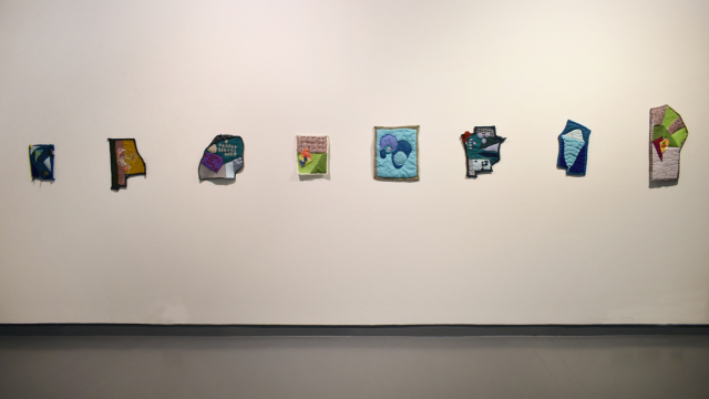 An exhibition photograph showing a row of small quilted pieces on the wall.