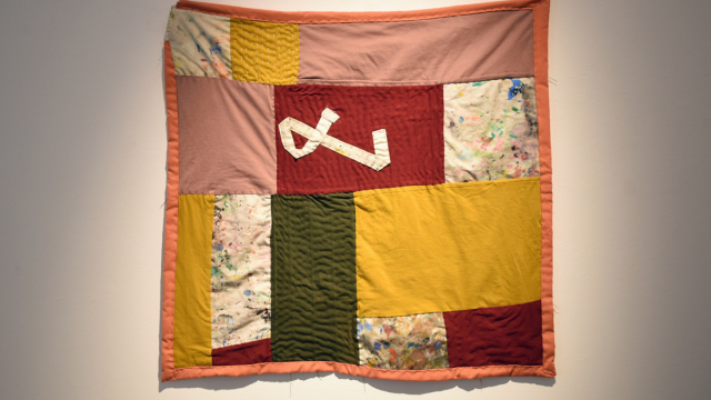 A wall quilt in ochre, dark green, coral, crimson and paint splattered fabrics, sewn with visible stitching and with a strip of white fabric sewn into a shape in the middle of the piece.