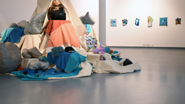 A photograph of the exhibition showing a fabric swaddled column with ledges of different heights - in the middle of the space. Perching on the various ledges and around the column are coloured fabrics, stuffed soft objects and shapes made with wool and fabric.