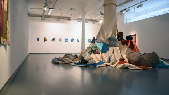 A photograph of the exhibition showing a fabric swaddled column with ledges of different heights - in the middle of the space. Perching on the various ledges and around the column are coloured fabrics, stuffed soft objects and shapes made with wool and fabric.