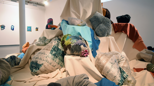 A photograph of the exhibition showing a fabric swaddled mountain shape with ledges of different heights - in the middle of the space. Perching on the various ledges are stuffed soft objects and shapes made with wool and fabric.