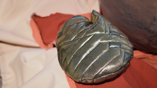 A round, shiny, silver metallic shape with quilted geometric lines sits on top of coral, brown and white fabrics which are out of focus in the background.