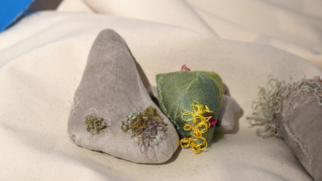 Angular stuffed shapes in silvers and greens are clustered together on a white canvas background. Looped embroidery in wool and threads covers parts of the objects, looking like moss or lichen.