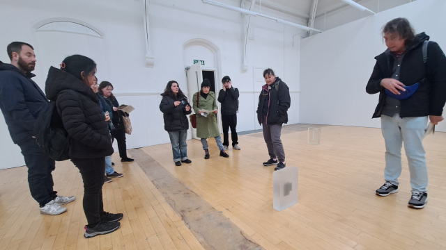 A group of people look at a small cube of resin resting on the floor. The resin holds an object inside it. They are all in a wide gallery space.