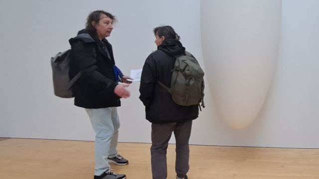 Two people stand beside a long oval shape in white. The shape is hanging from the wall in a gallery space.
