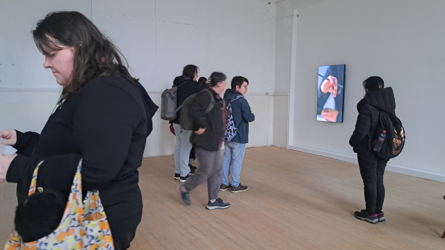 A group of people in a gallery space. There is a screen showing someone's hands on the far wall.