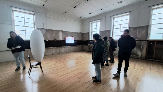 A spacious gallery with wooden floors. There is a 3 dimensional oval shape on top of a wood chair and a group of people looking at it.