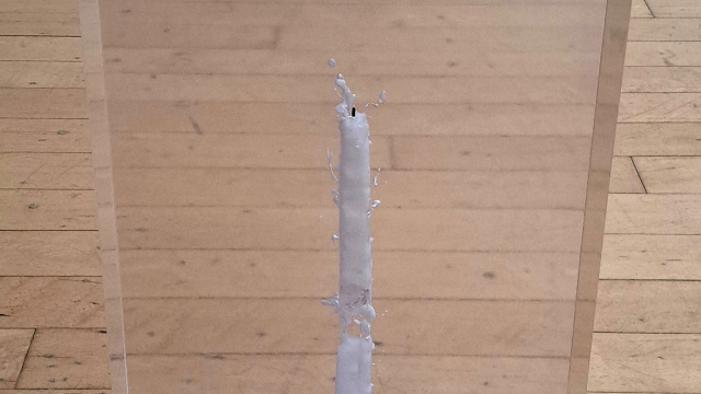 A candle inside a block of clear resin. The candle is broken. The block of resin is on the floor in a gallery space.