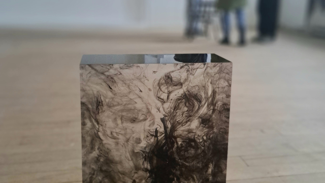 A block of clear resin with a pen and swirls of ink inside it. It is on the floor in a gallery space.