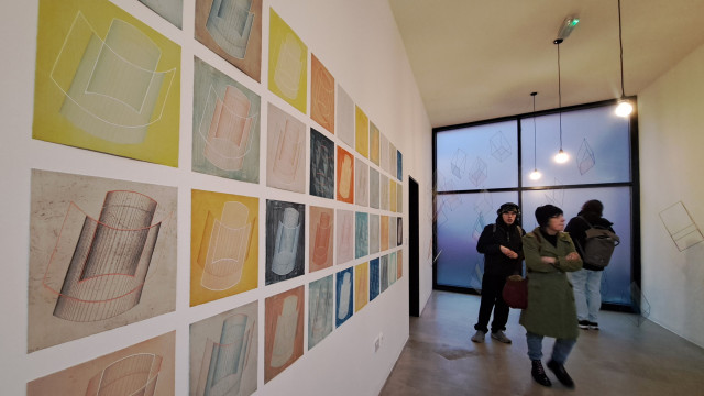 Three people in a narrow gallery space. The wall on the left has 4 rows of the same art print in different colour combinations.