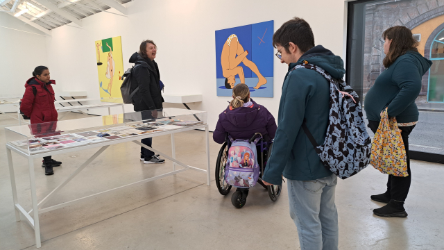 A group of people gather inside a spacious gallery, there are big artworks on the walls and more vitrines in the open space.