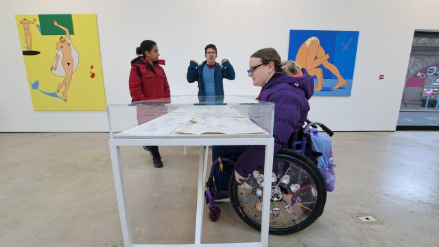 A wheelchair user inspects a vitrine in a gallery space. There are big artworks hung on the walls on the back and two people stand further away.