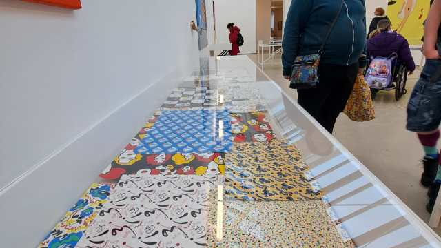 A long vitrine within a gallery space, with many artworks inside it. The artworks have a variety of patterns with bright colours on them.