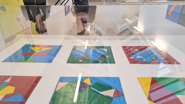 6 bright square artworks inside of a glass vitrine.