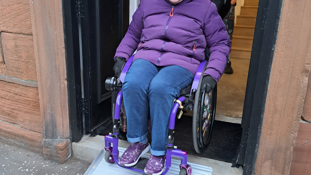 A wheelchair user is exiting a gallery space using a ramp. Someone is helping them out.