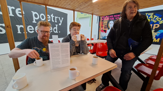 Two people enjoy a cup of tea in a pop up café space inside a gallery. Another person is perching on the table next to them.