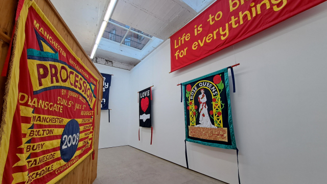A photo of fabric banners in a gallery space. The banners are made in bold colours. On of them has the words "Life is to blame for everything" sewn on it.