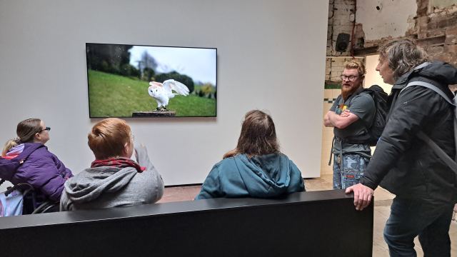 People sit on and stand around a bench, looking at a screen. There is a barn owl on the screen.