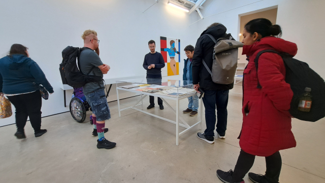 A group of people in a gallery space gather around a vitrine with several artworks.