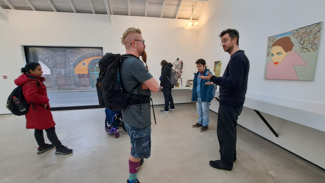 A group of people stand in a gallery space, the two closest to camera are chatting to each other.