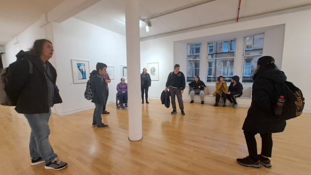 A group of people in a gallery space, some are standing up and looking around while other take a sit and talk to each other.
