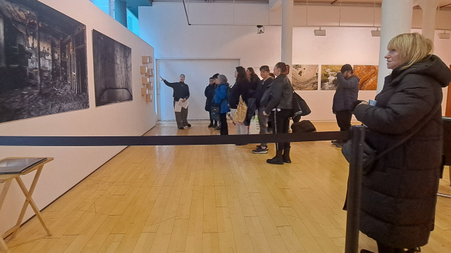 A group of people look at artwork in a spacious gallery. Someone is pointing at the art. There is a person standing closer to the camera.