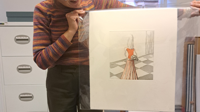 A person holds up a print of a woman with her head covered by a white cloth. The woman in the print is holding her own head in a serving tray.